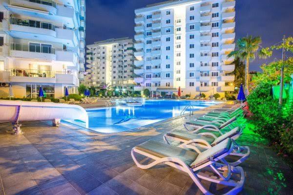 Gece ışıklandırılmış açık havuzda gri şezlonglar ve mavi kapalı şemsiyeler, satıştaki sitede