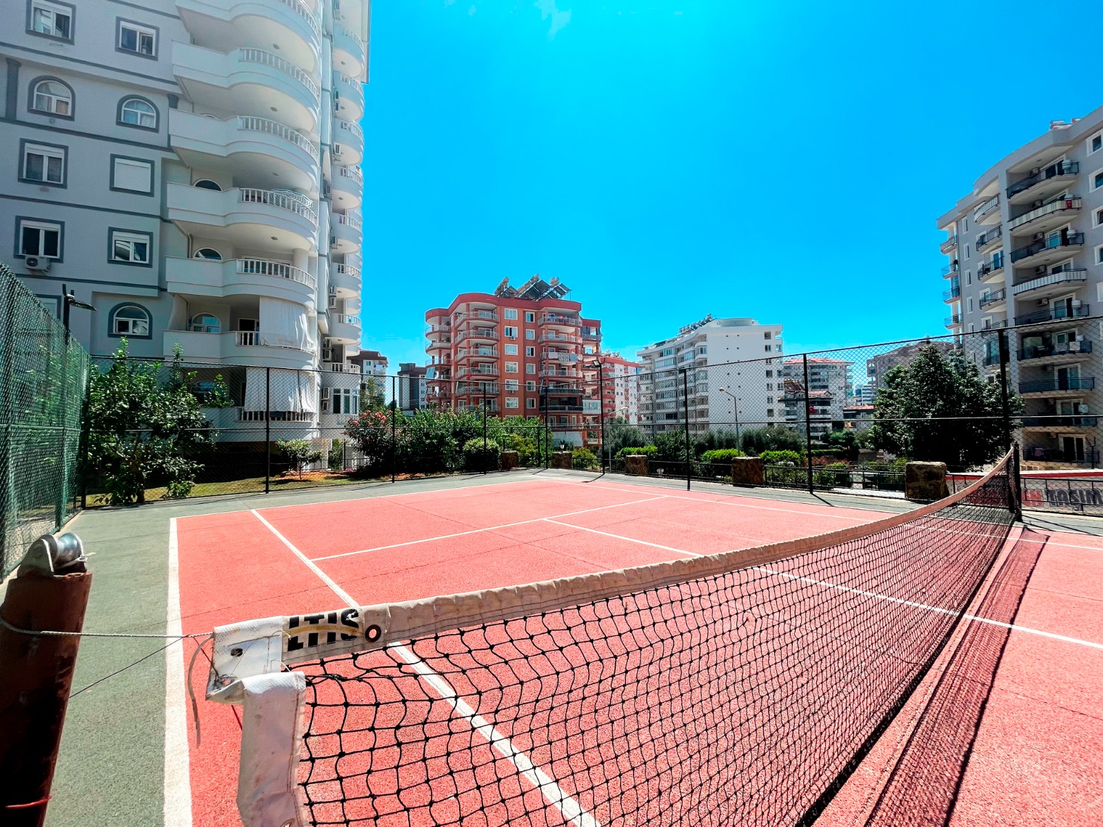 Ön planda tenis ağı ve yüksek tel çitli kırmızı spor kortu, denize yakın sitede