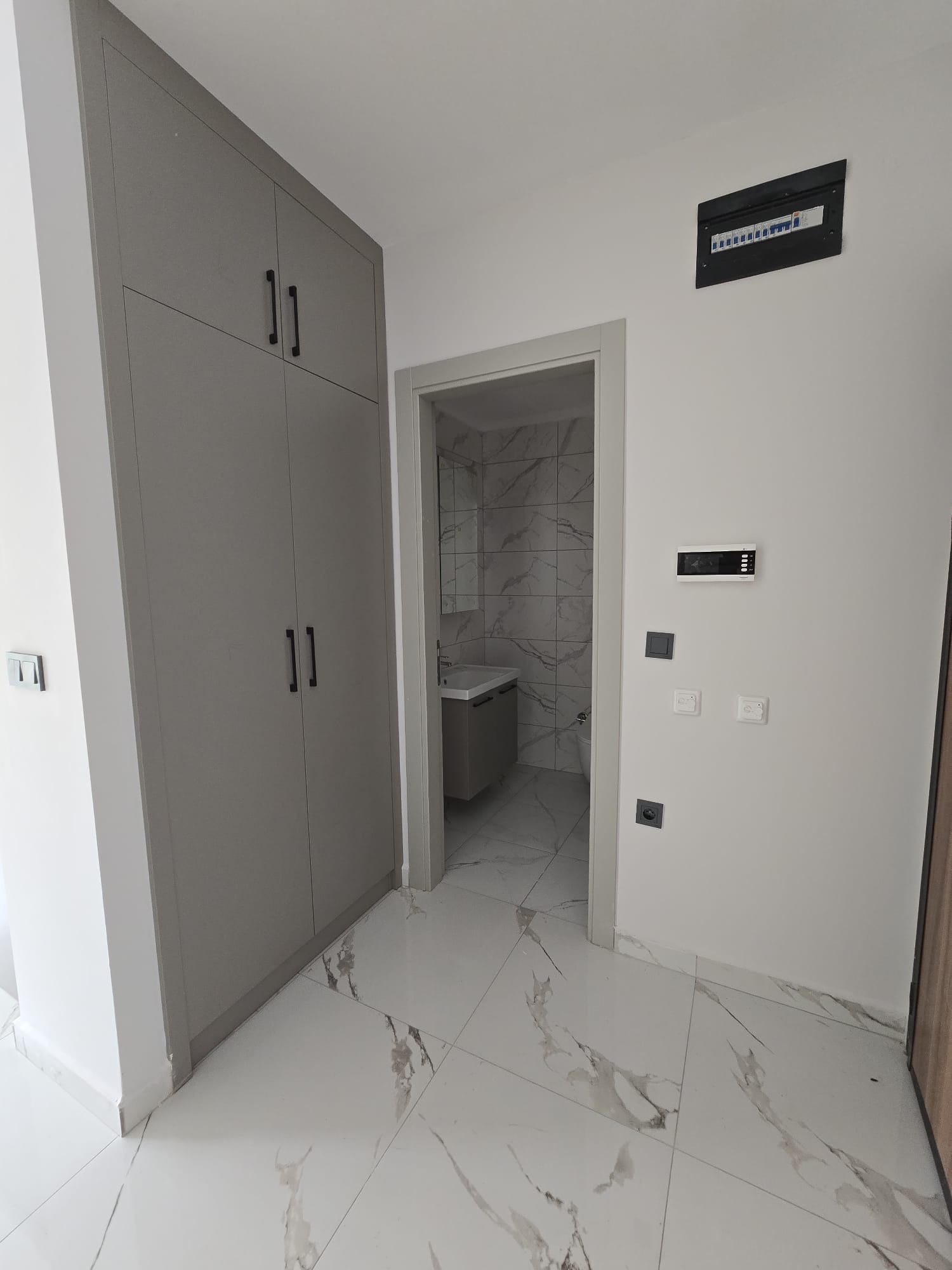 Gömme yüksek dolaplı antrede banyo kapısı, elektrik panosu ve mermer desenli zemin konutta