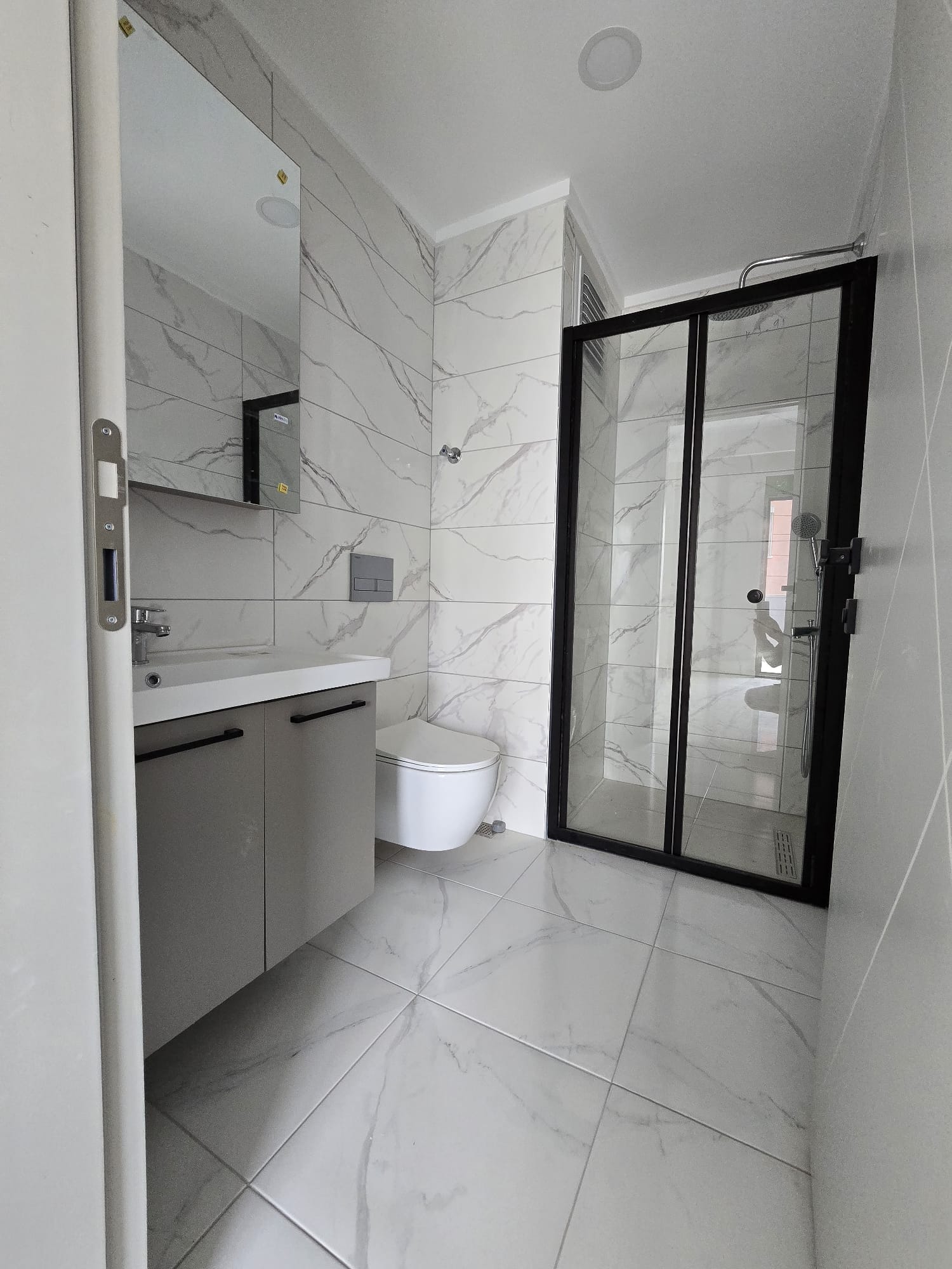 Siyah çerçeveli cam duşlu sıhhi hacimde gri dolaplı lavabo ve mermer fayans Tosmur’daki konutta