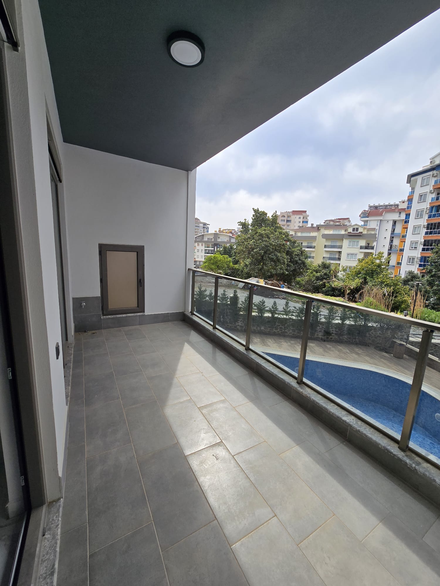 Kapalı terasta cam korkuluk, havuz ve ağaçlı avlu manzarası satılık evde