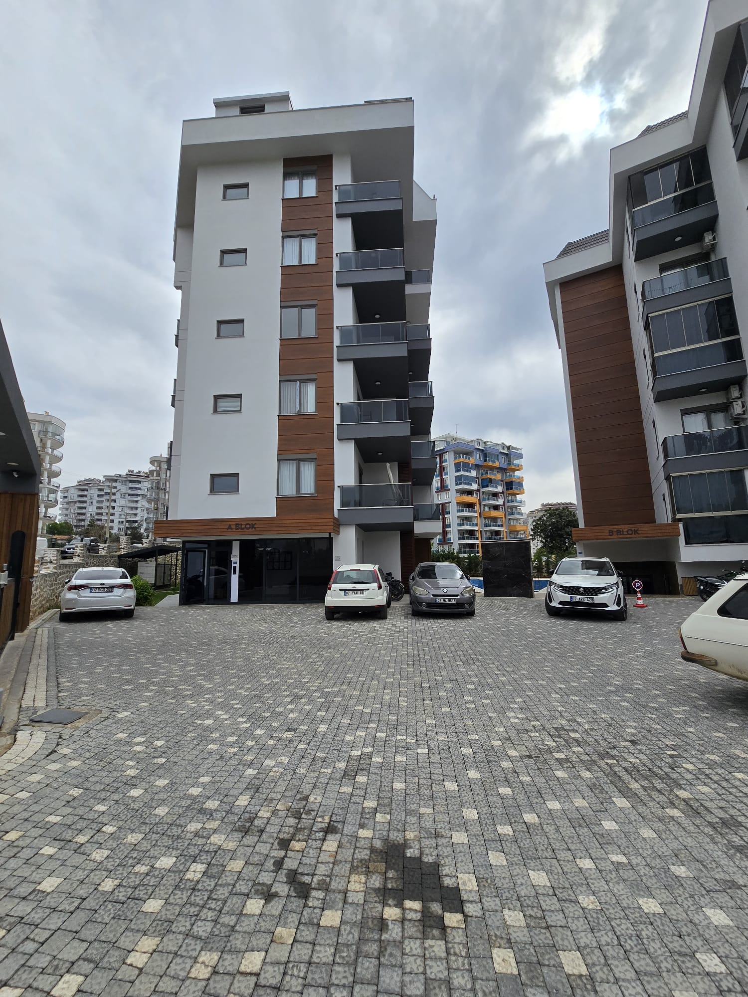 İç otoparkta gri taş döşeme, balkonlu bloklar ve şehir yapıları Tosmur’daki dairelerin bulunduğu sitede