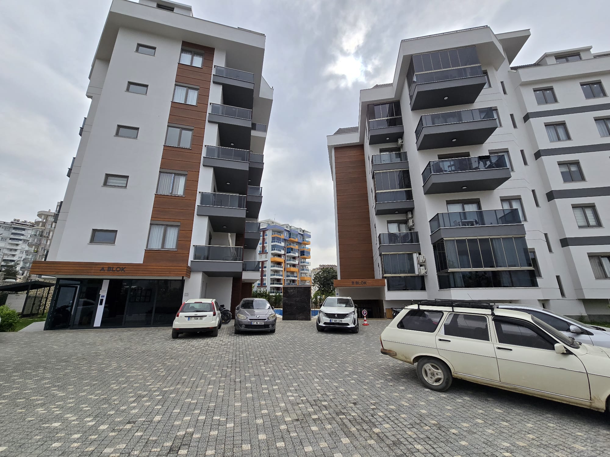 Bloklar arasındaki gri taş döşeli otopark, cam balkonlar ve araçlar Tosmur’daki konutların bulunduğu sitede