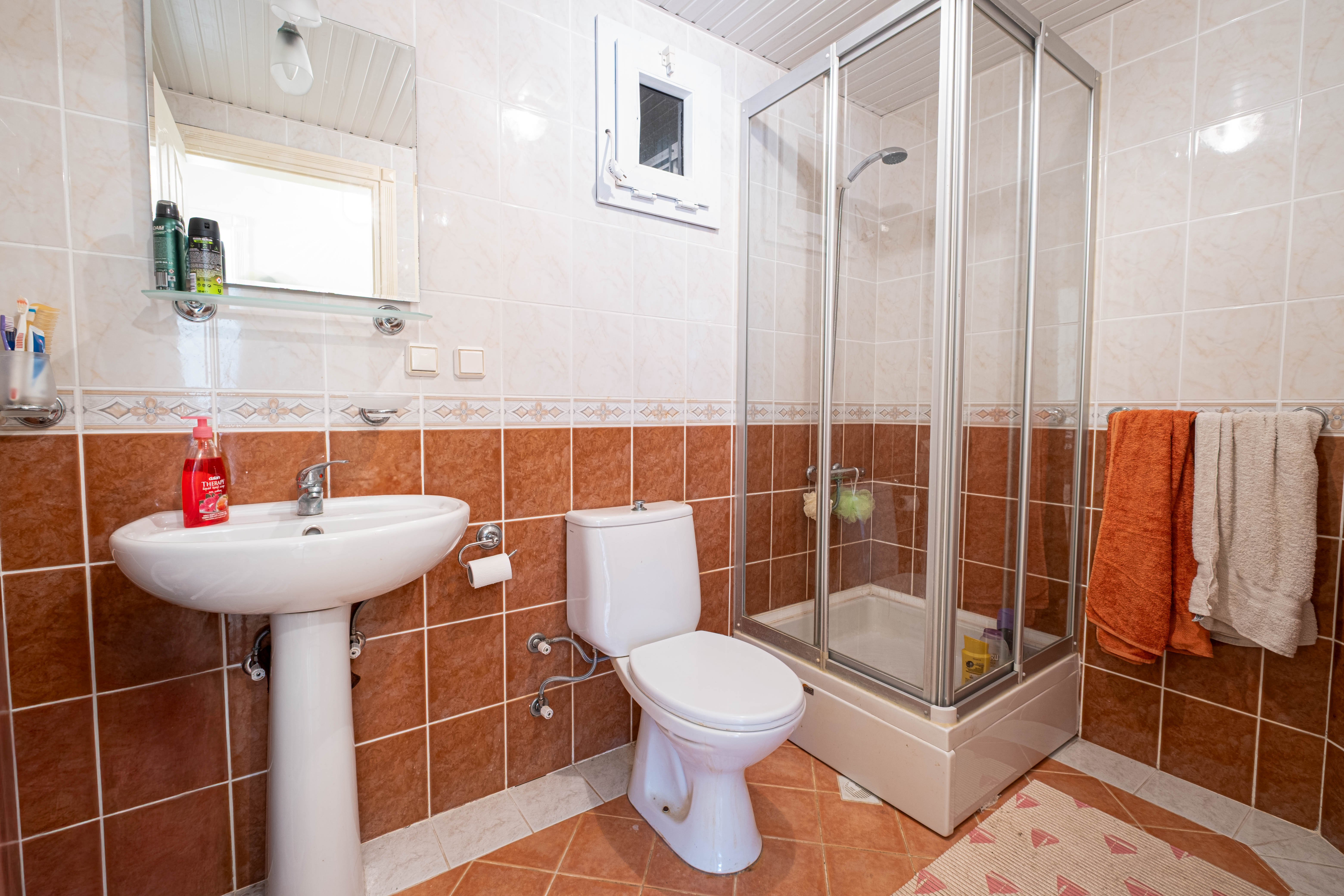 Banyoda cam duş kabini kaideli beyaz lavabo duvar aynası ve dekoratif bordürlü seramikler bulunuyor