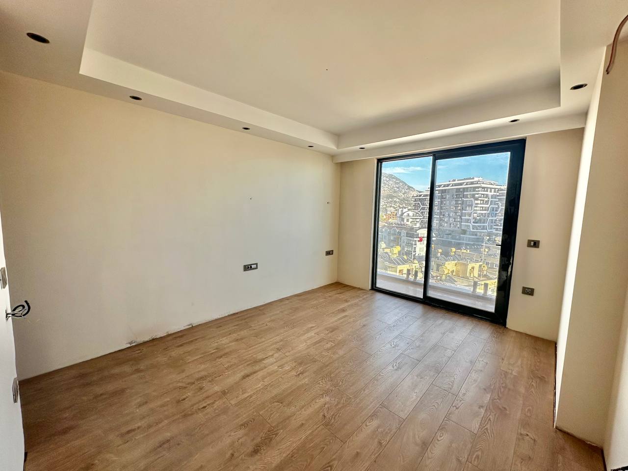 Ahşap zeminli boş yatak odası, Tosmur’da panoramik pencereden şehir binalarını ve dağ yamacını gösteriyor