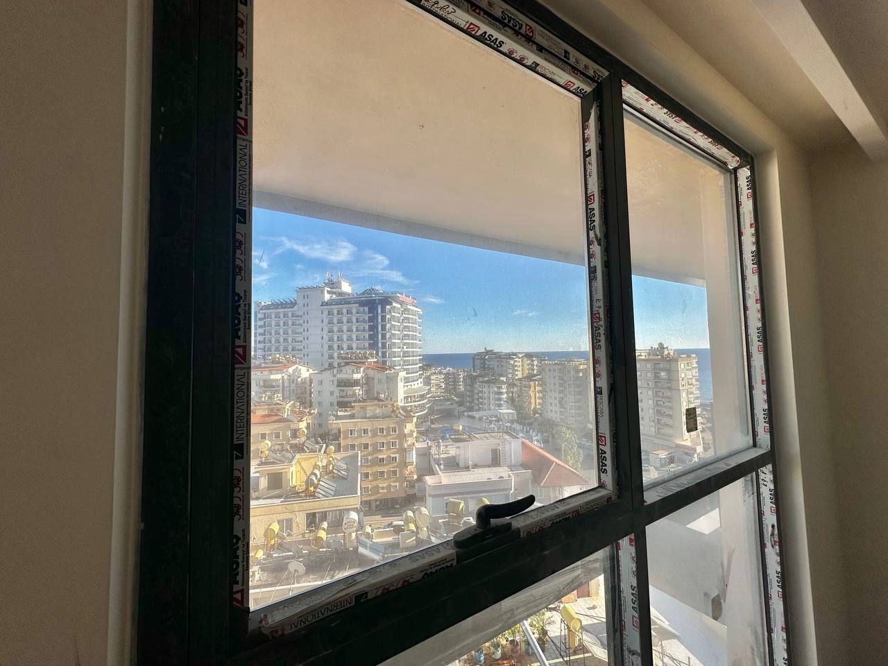 Camdan görünen panoramik şehir manzarasında uzakta mavi deniz şeridi ve çok katlı binalar yer alıyor
