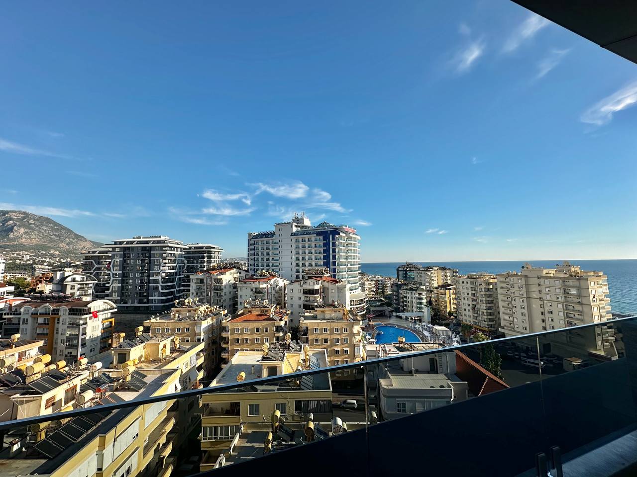 Terasın cam korkuluğundan deniz, dağ yamacı ve avludaki yuvarlak havuz görülüyor