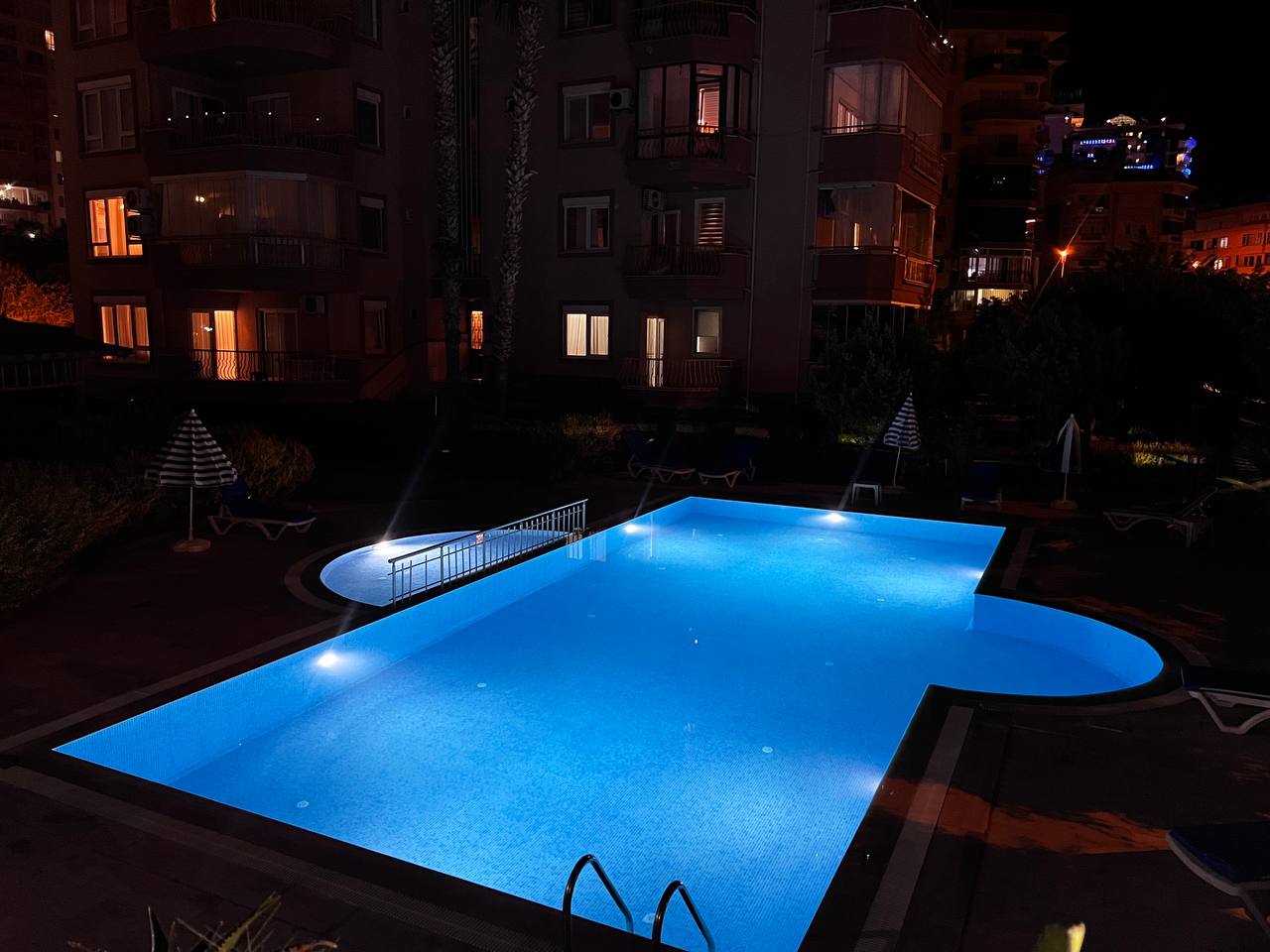 Illuminated outdoor pool with blue water and loungers in a complex with residences in Tosmur, Turkey