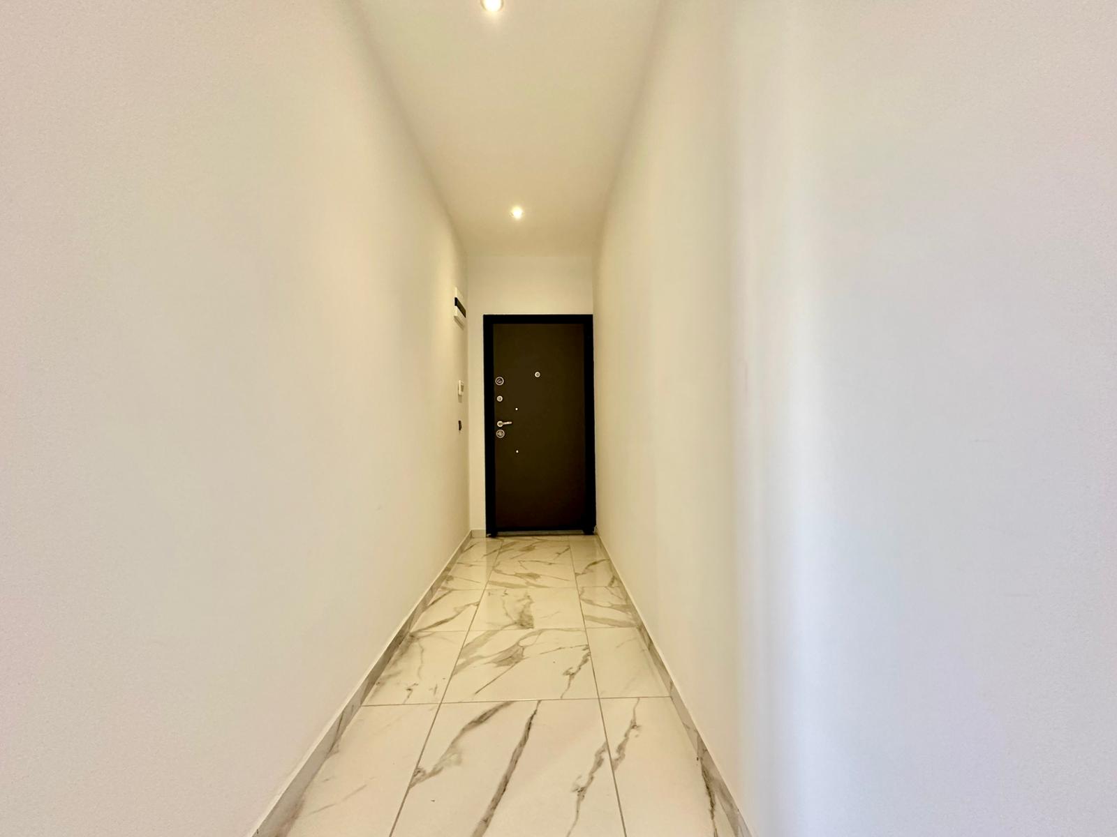 Dairenin koridorunda mermer desenli açık renk karolar ve uçta koyu renk giriş kapısı